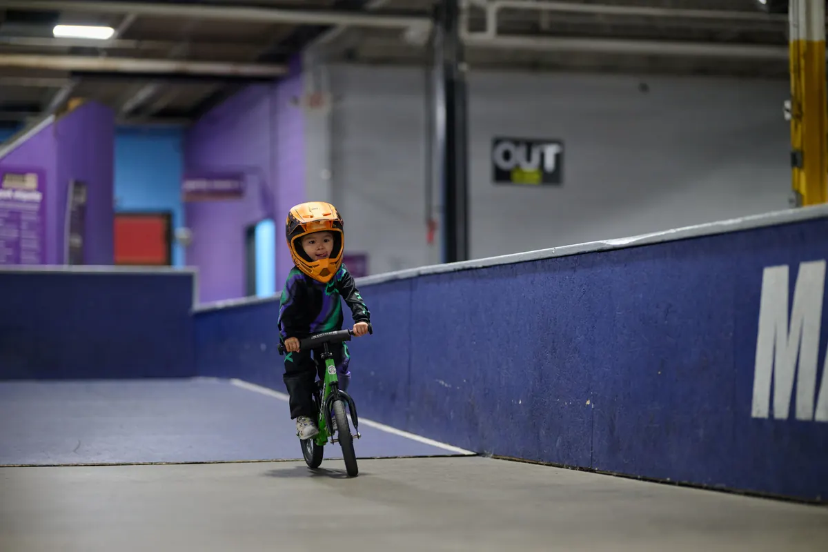 A young child learning to ride a balance bike at an indoor XKIDS practice space.